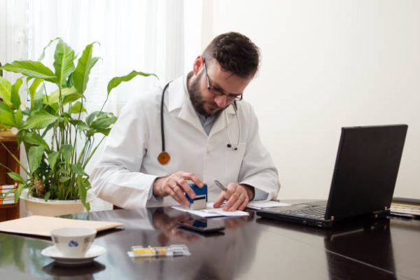 A doctor in a white coat sitting at the table and affix a stamp to the document. Doctor's hand stamped on the prescription. The doctor writes a prescription at his desk. The doctor sits at a table and signed prescription issued.